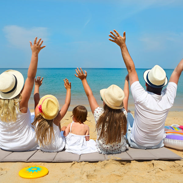 Family of five with hats sitting on a beach mat, raising their hands towards the sky.