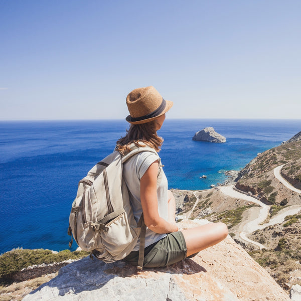 Person with a backpack and hat sitting on a cliff overlooking the ocean.